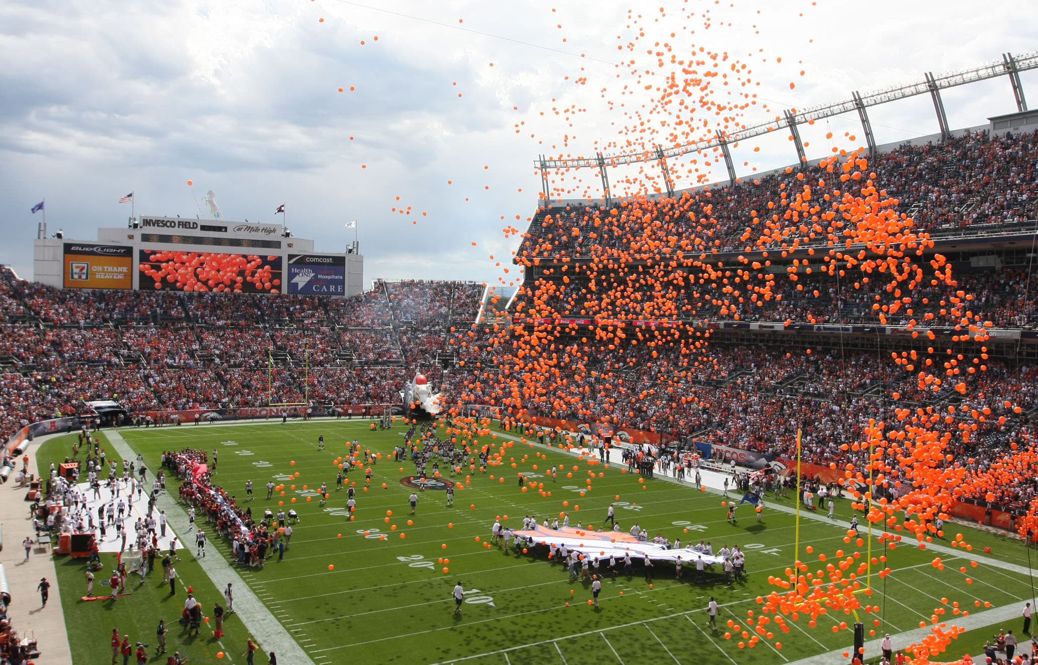 Orange balloons float through the air at the Denver Bronco's football stadium. People on the field hold out a giant Broncos logo.