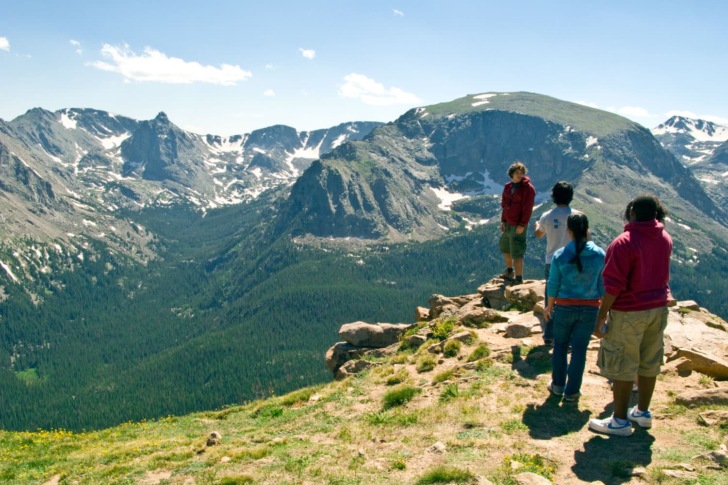 A group of teens stands around a rocky scenic overlook to admire the expansive, lush forested valley below and majestic mountains with pockets of spring snow in Colorado.