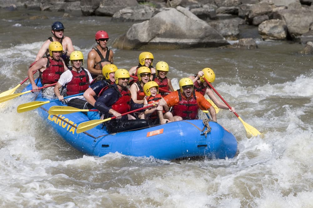 12 people in yellow helmets and red life jackets hold yellow paddles and ride the river waves in an inflatable blue boat