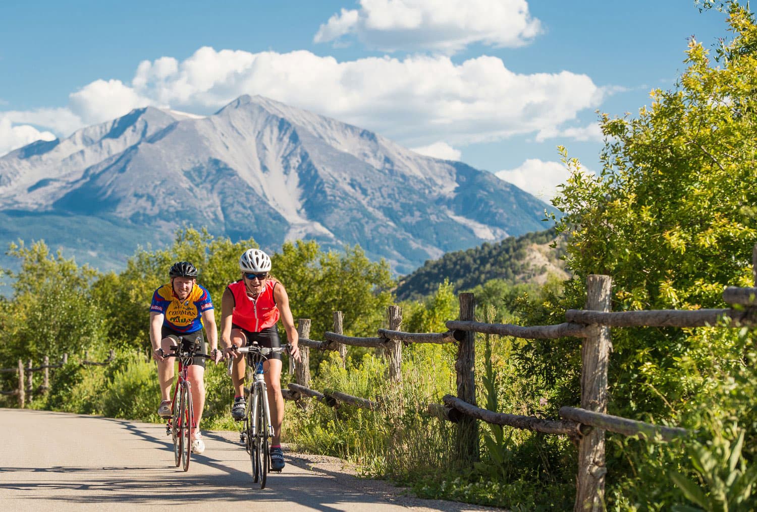 Two cyclists road bike next to simple, wood-post fence in Glenwood Springs, Colorado. The trees along the fence a vibrant with summer color and the mountains behind them are capped with snow.