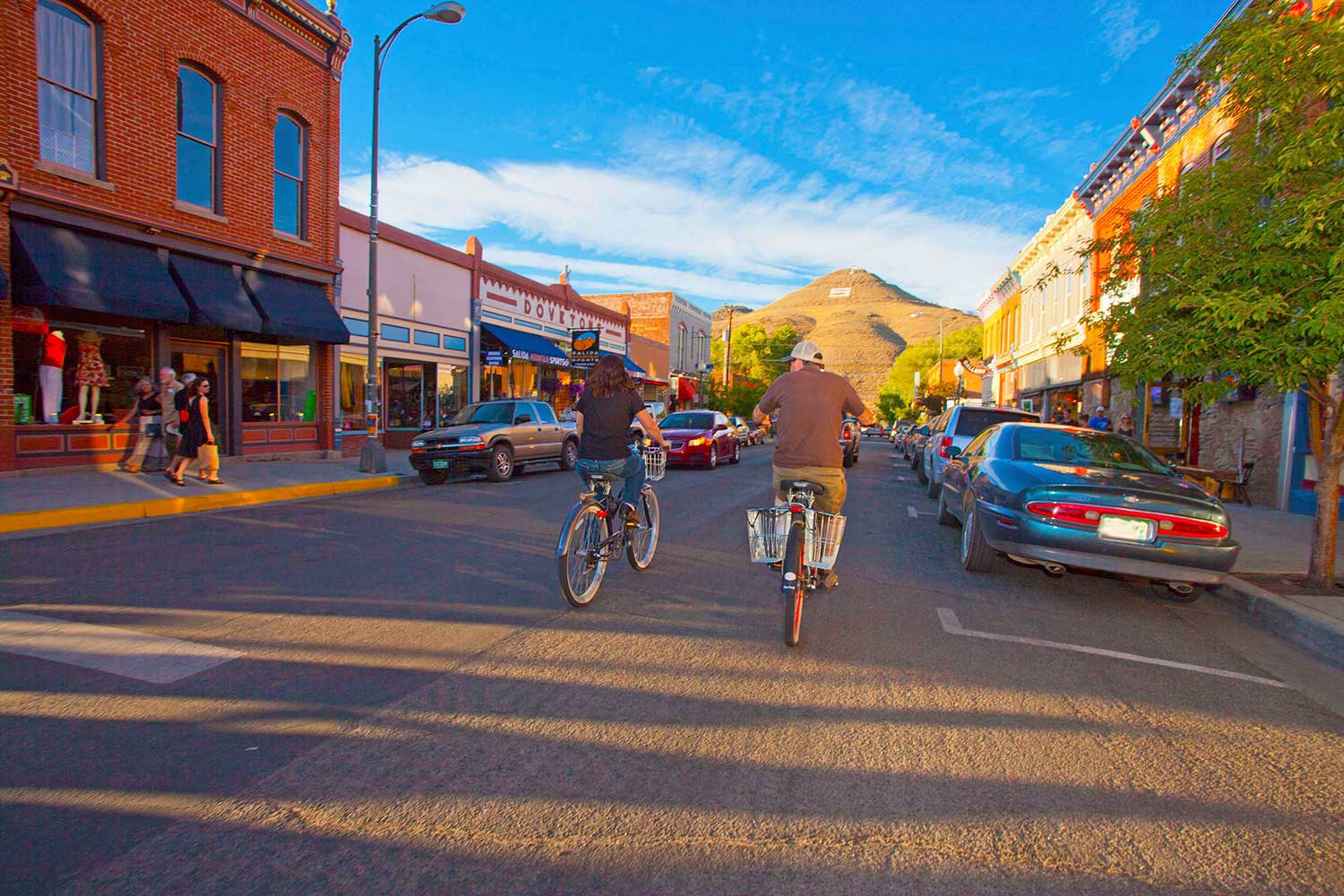 Two people ride cruiser bikes along a street lined with cars and pedestrians walking by shops. The town's "S" for Salida can be seen chalked in white on the side of hill at the end of the street