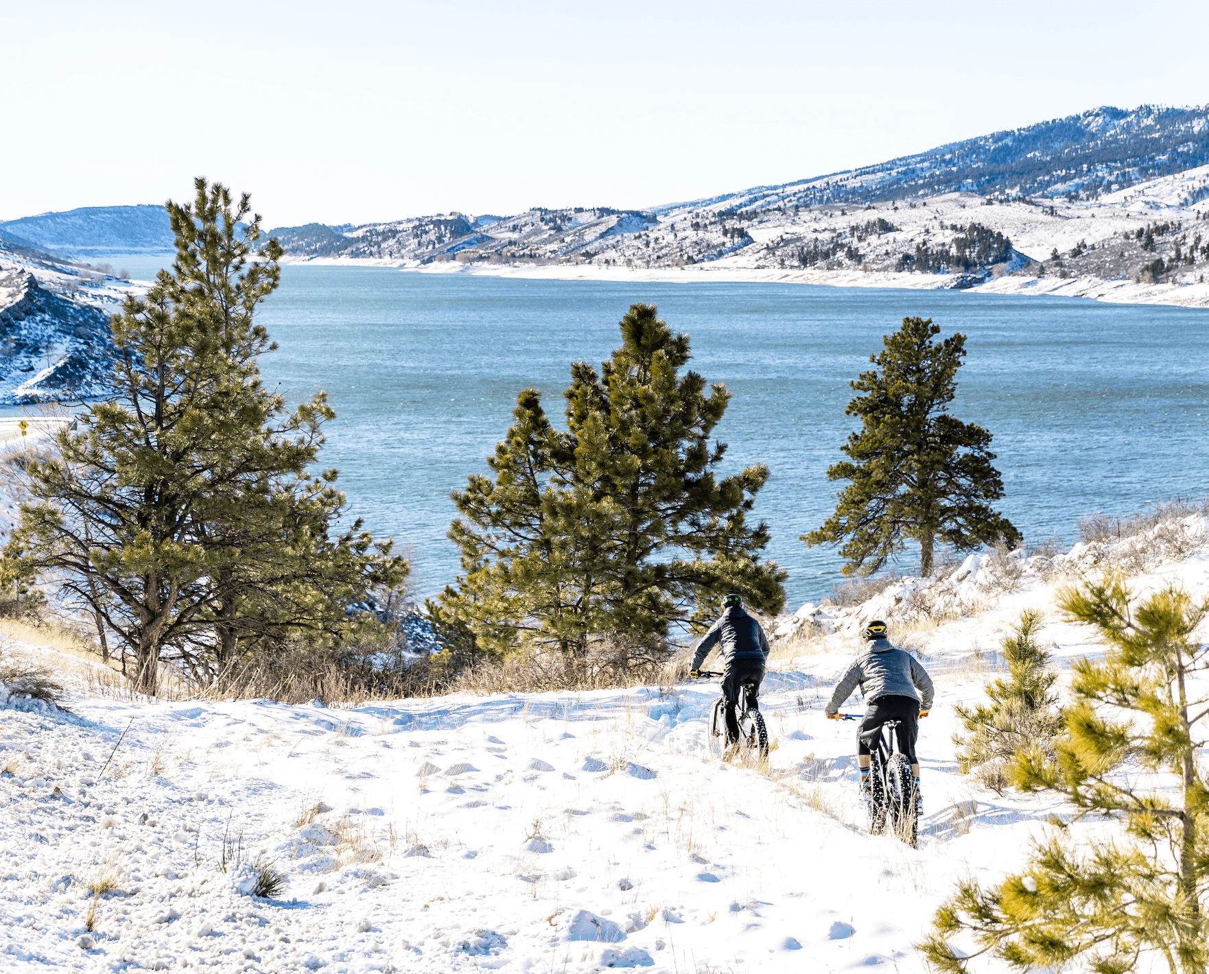 Two people ride fat bikes on a snowy trail overlooking a blue reservoir with evergreen trees in Fort Collins, Colorado.
