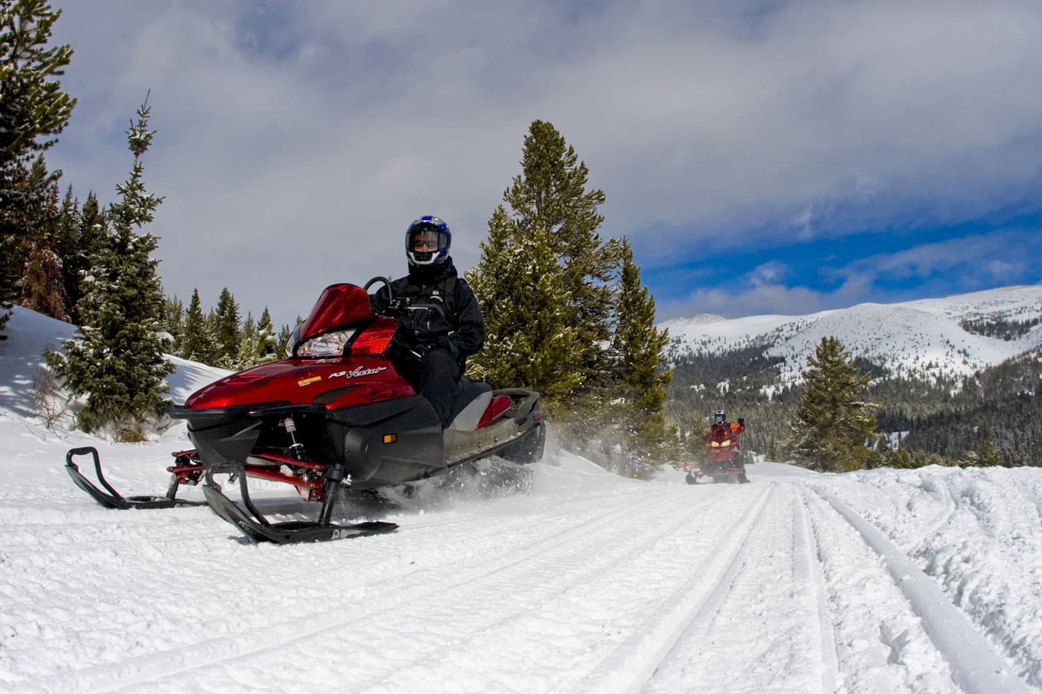 A snowmobile glides across a snowy trail with trees and mountains in the background