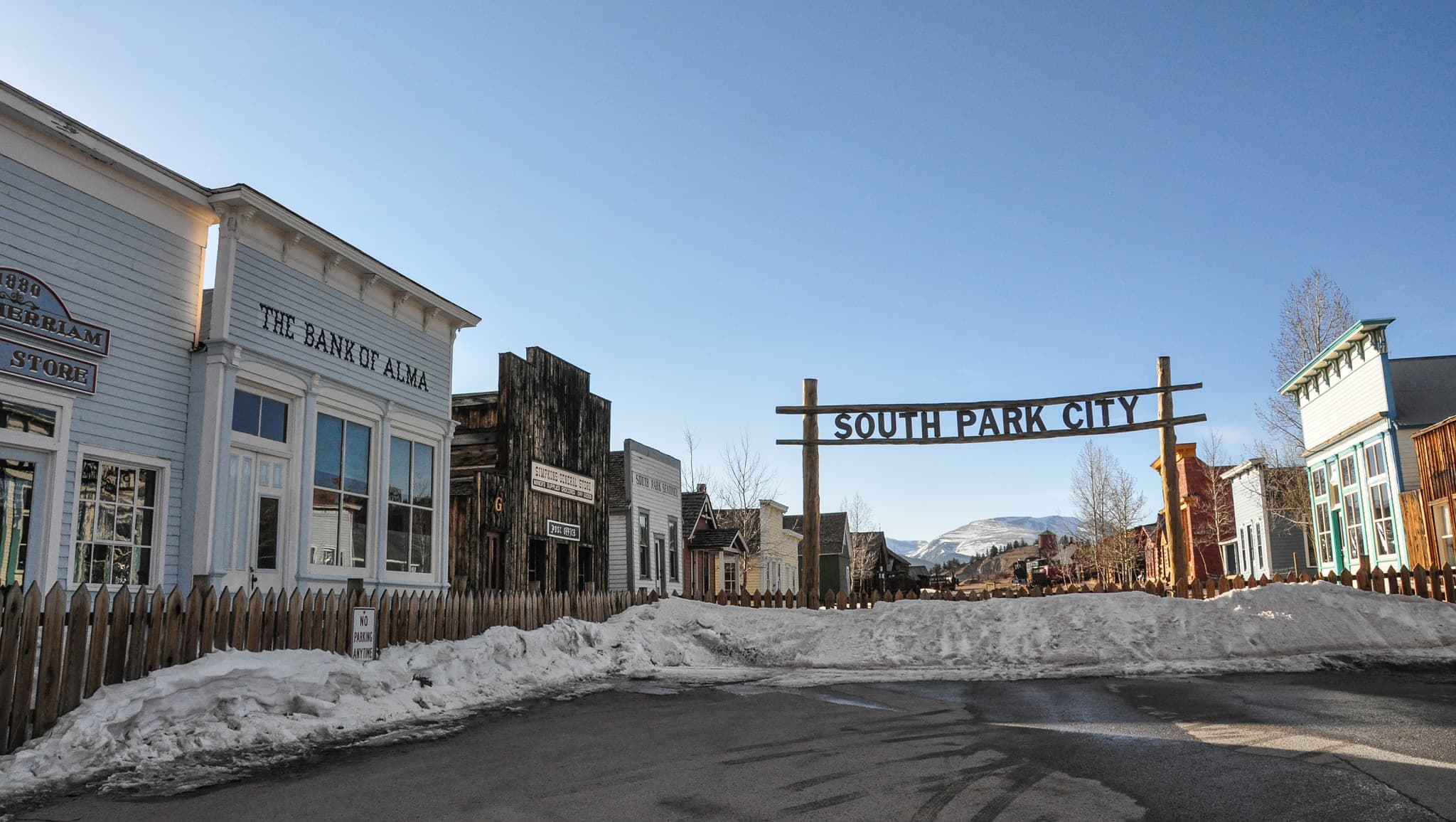 Snow is piled up along a low, plain wooden picket fence to reveal a clear black top area in Colorado. A nearby wooden sign reads, "South Park City."