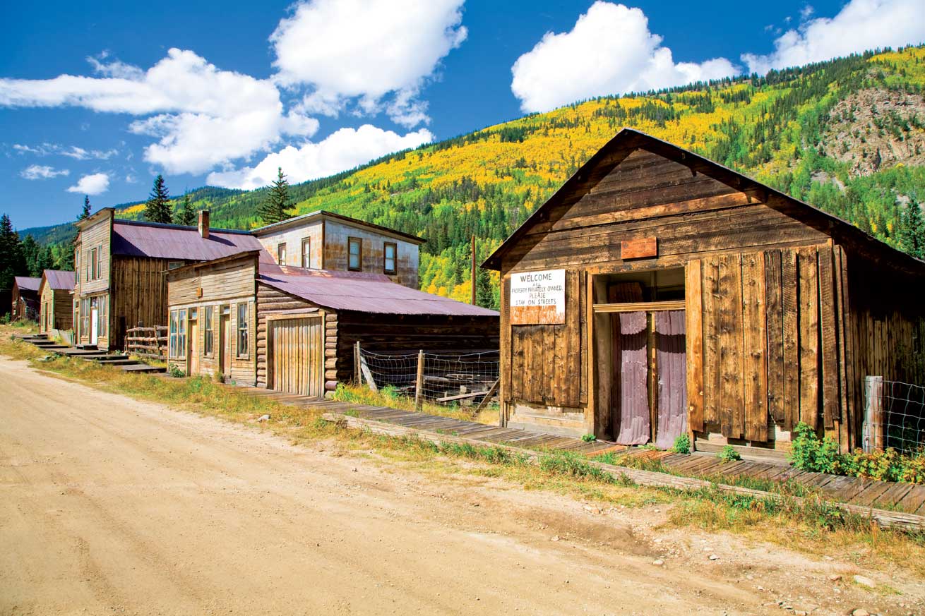 Abandoned wood buildings show signs of disrepair and stand empty along the side of a dirt road near Buena Vista. Behind them, trees begin showing their golden fall colors.