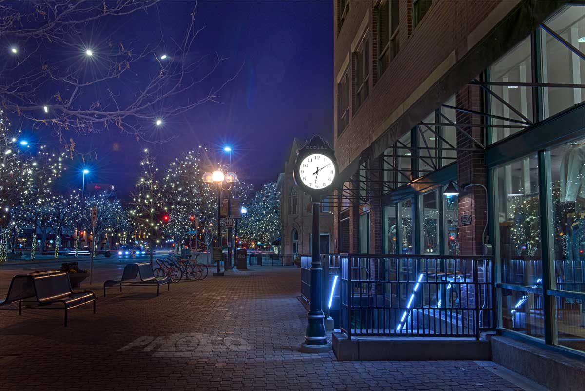 The streets of Fort Collins, Colorado, are empty on an early winter morning. A street clock and lights on trees down the road glow bright in the twilight.