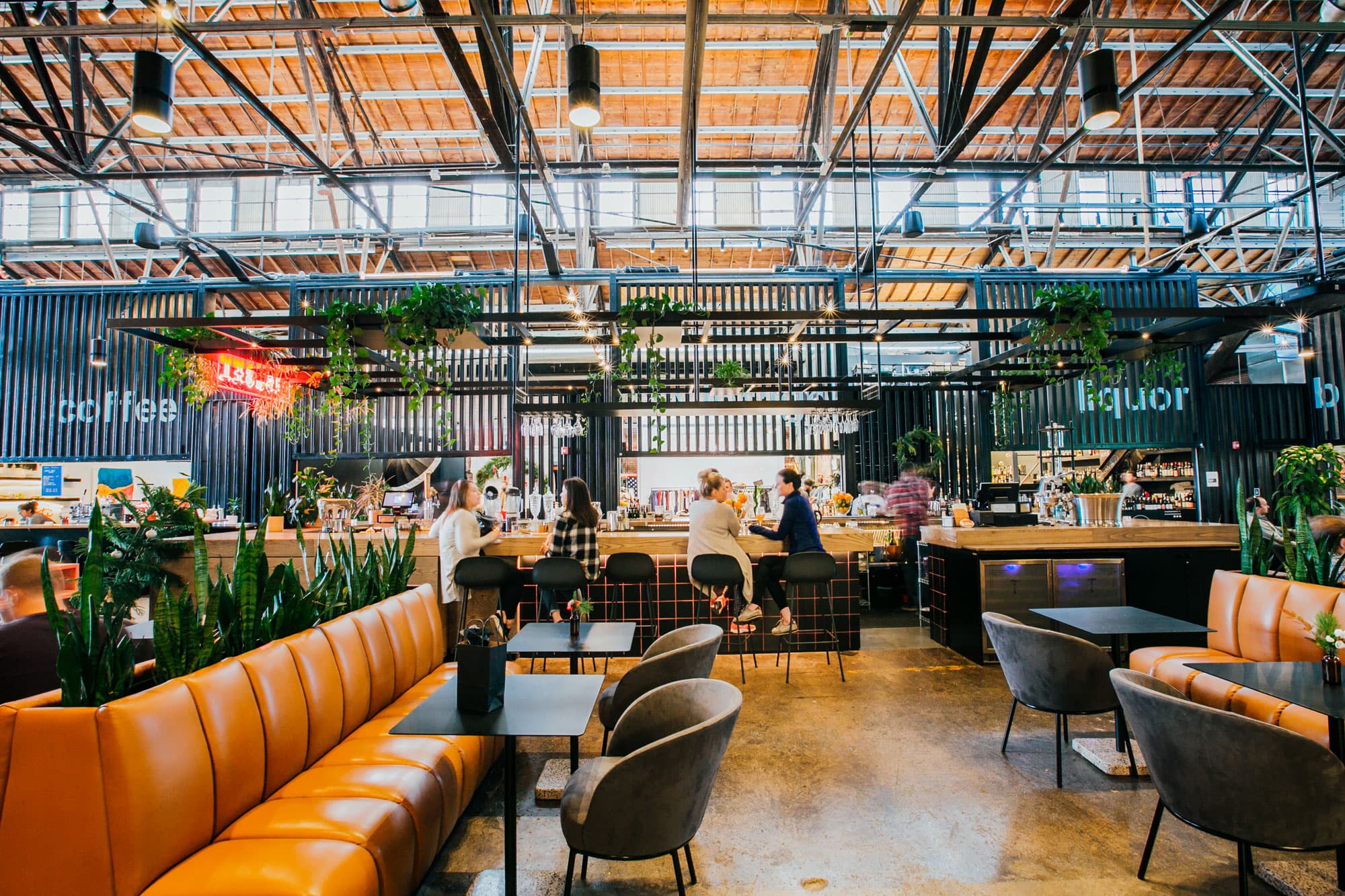 Four people sit in black bar stools at a wood-top bar at a market hall in Denver, Colorado. The space is decorated with leather benches, snake plants and dripping pathos plants.