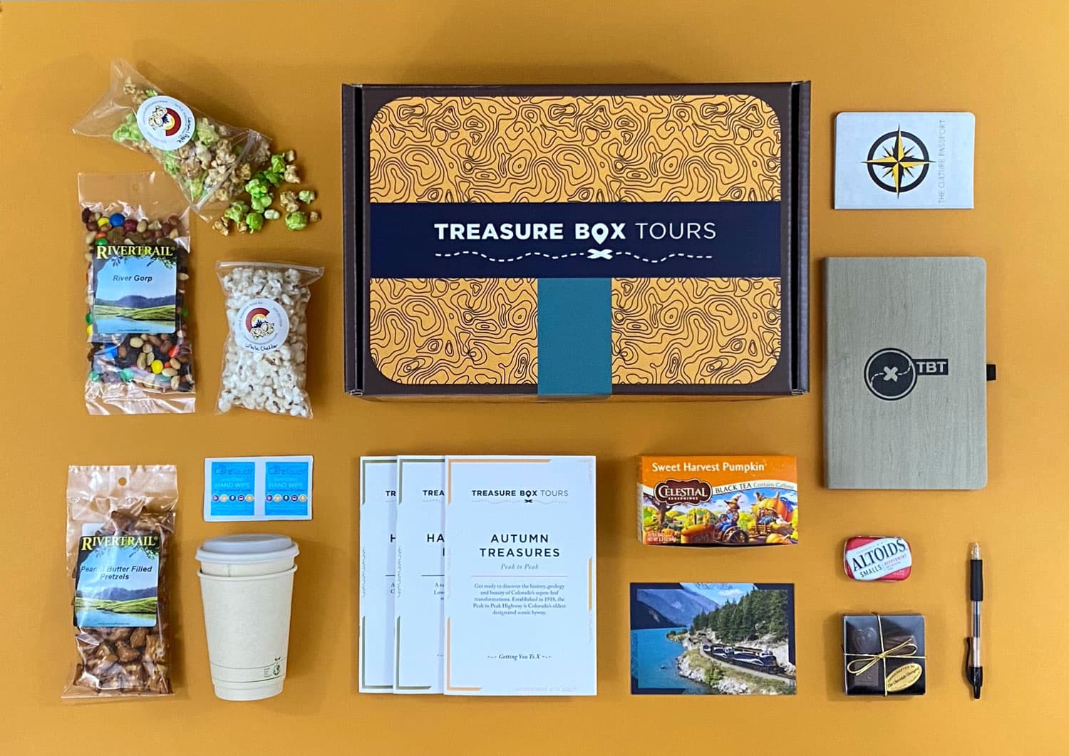 Several items are laid out flat on a burnt-orange background. The items include several popped popcorn and trail mix packages, a box of tea, a black pen and a gray, fabric-covered notebook.