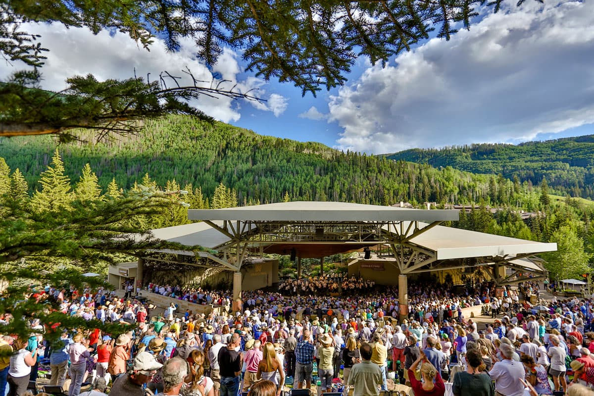 A crowd lined up in front of an amphitheater with evergreen forest behind it