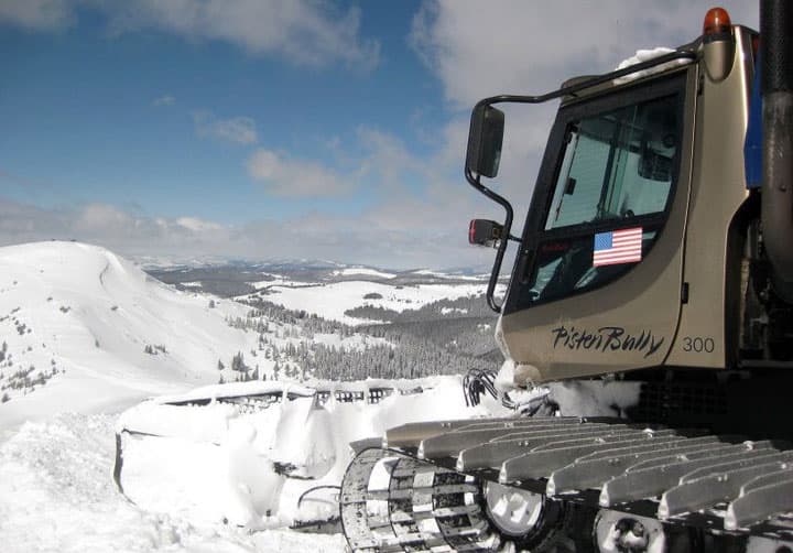 A closeup of a gray snowcat parked near the top of a mountain in Colorado reveals a snow-packed front bumper and deep-tread wheels.