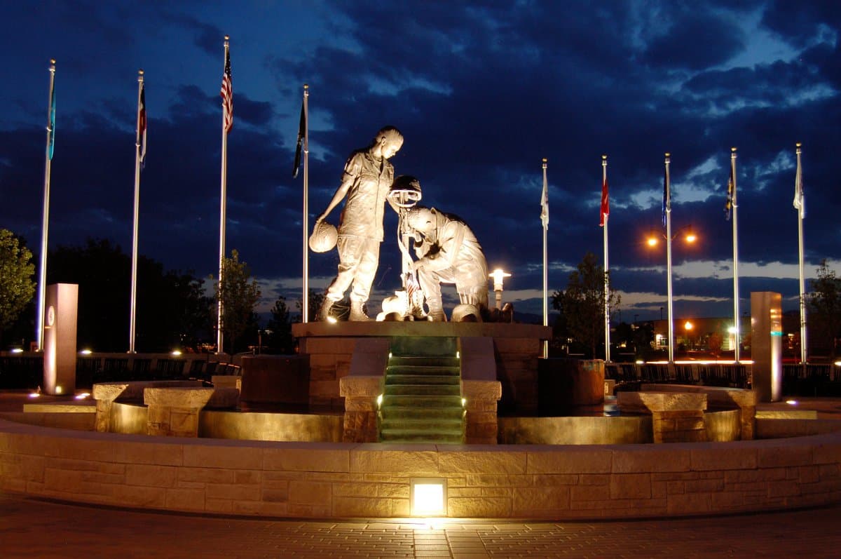 An nighttime viewing of the Armed Forces Tribute Garden in Westminster. In the middle a statue of a soldier down on one knee with another soldier standing over him. In the background there are flag poles.