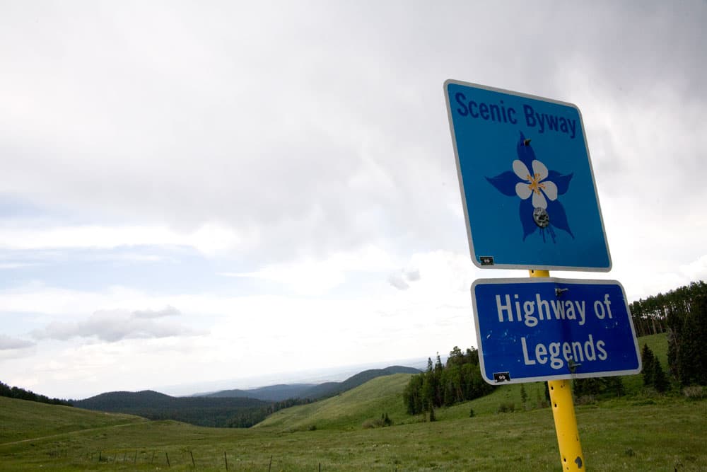 A green, mountain vista along the Highway of Legends Scenic and Historic Byway in southeast Colorado. On the right a metal square sign with a flower on it says "Scenic Byway." Below that on the yellow sign pole is a blue rectangular sign that says "Highway of Legends."