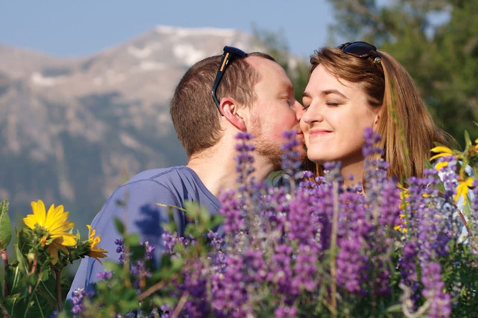 A person kisses their smiling partner's cheek as the two sit amid a field of blooming purple and yellow wildflowers in Colorado.