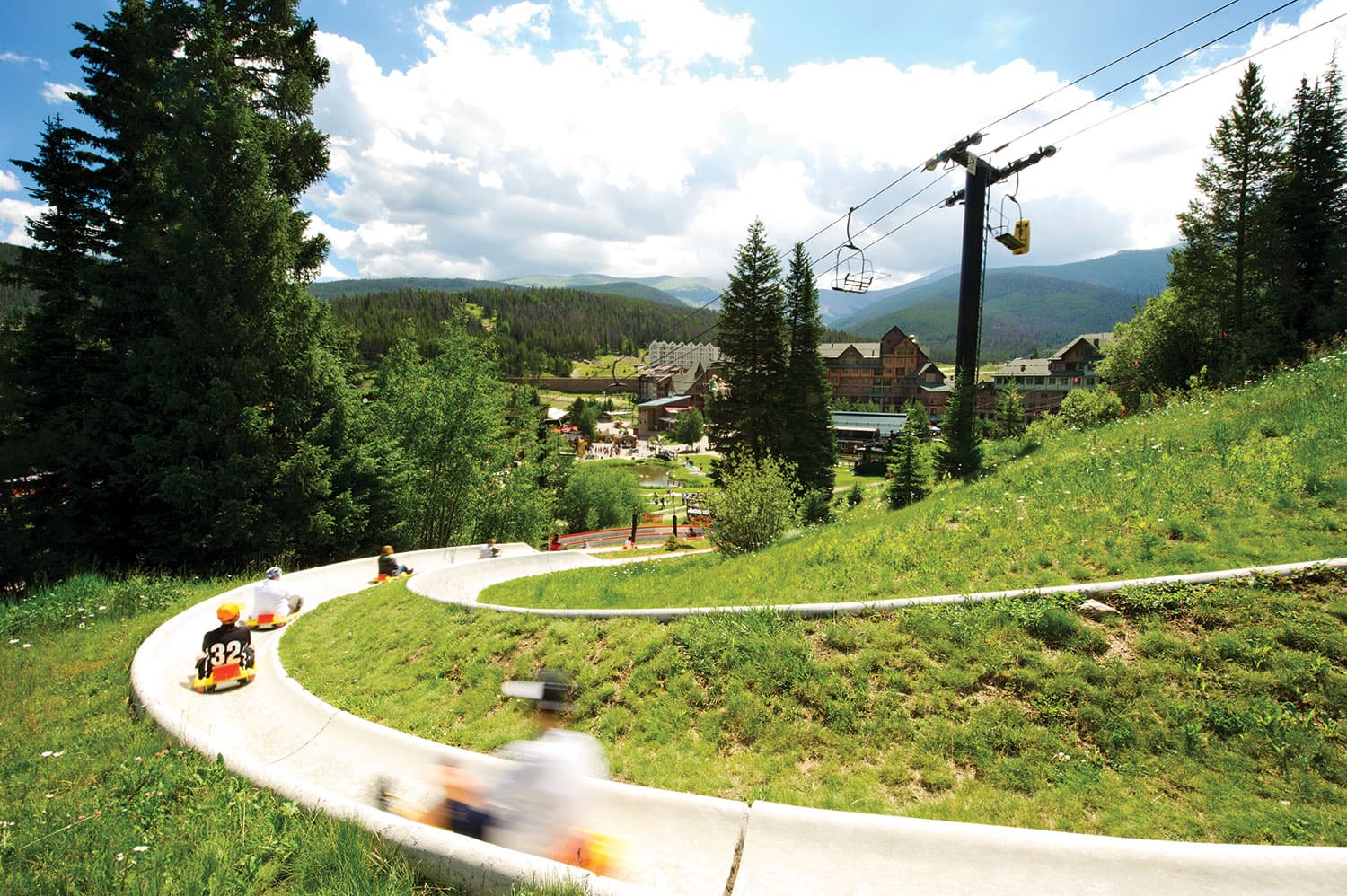People sitting on yellow mats fly down a white half-tube slide that curves down a steep mountainside at Winter Park Resort in Colorado.
