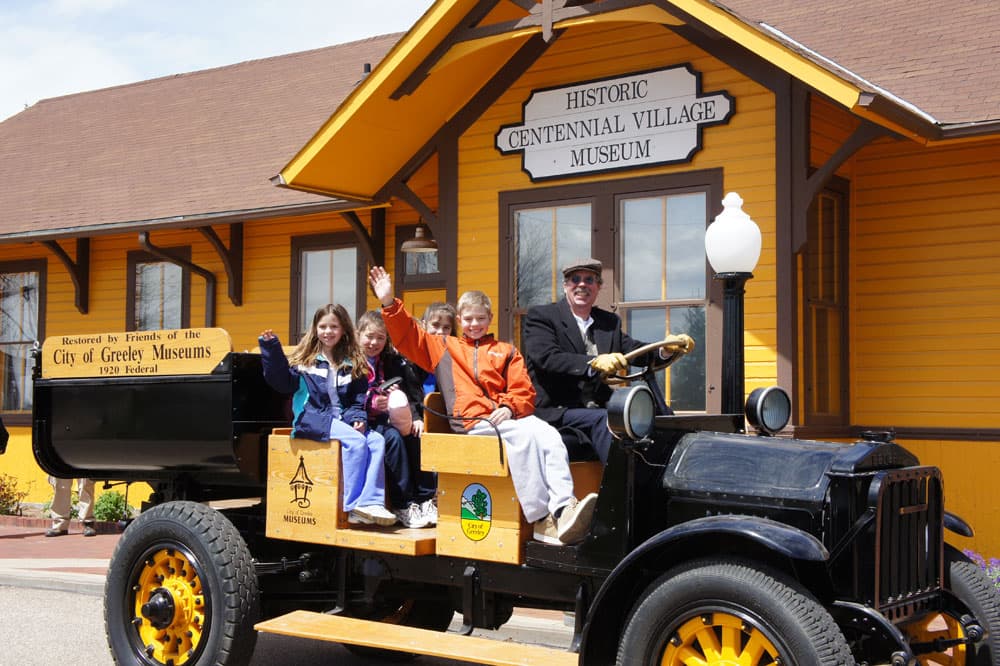 In front of the yellow wooden building with brown trim that has a sign that reads "Historic Centennial Village Museum" sits an old truck being driven by a man. Three children it in the seats, one waves at the camera while wearing an orange jacket.