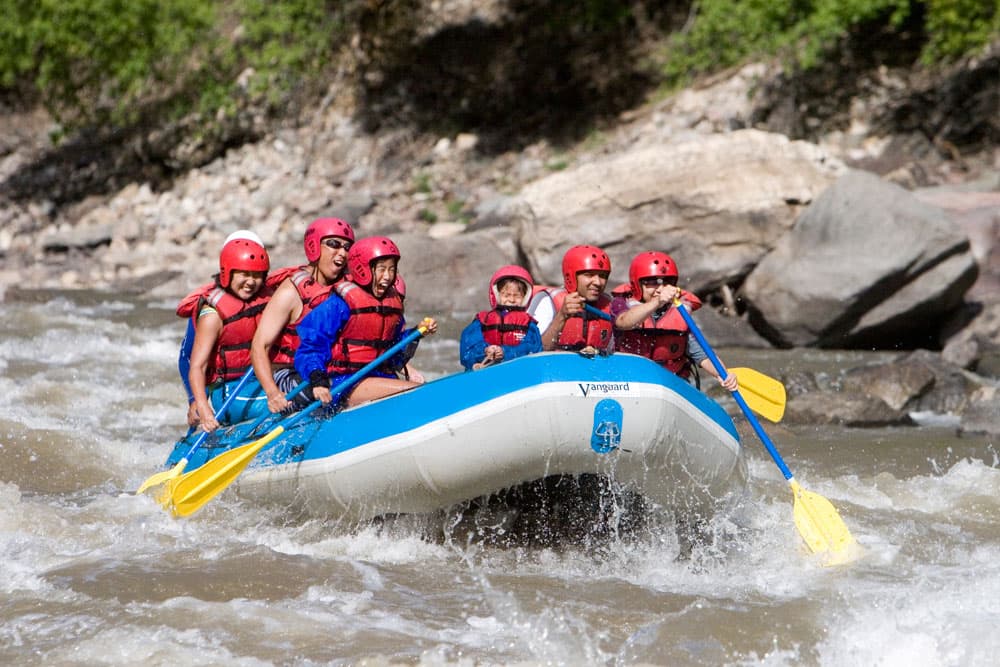 A blue and great boat tips its nose up as it crests a wave. Inside, paddlers in red helmets and life jackets grip their paddles as they wait for the next one