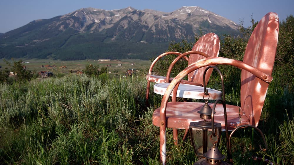 Two white but rusted-over metal lawn chairs sit in a field of tall, green grass. Between them is a small round, white metal table, also rusting but not as much as the chairs. In the background is a blue-and-gray mountain range underneath a dusty-blue sky.