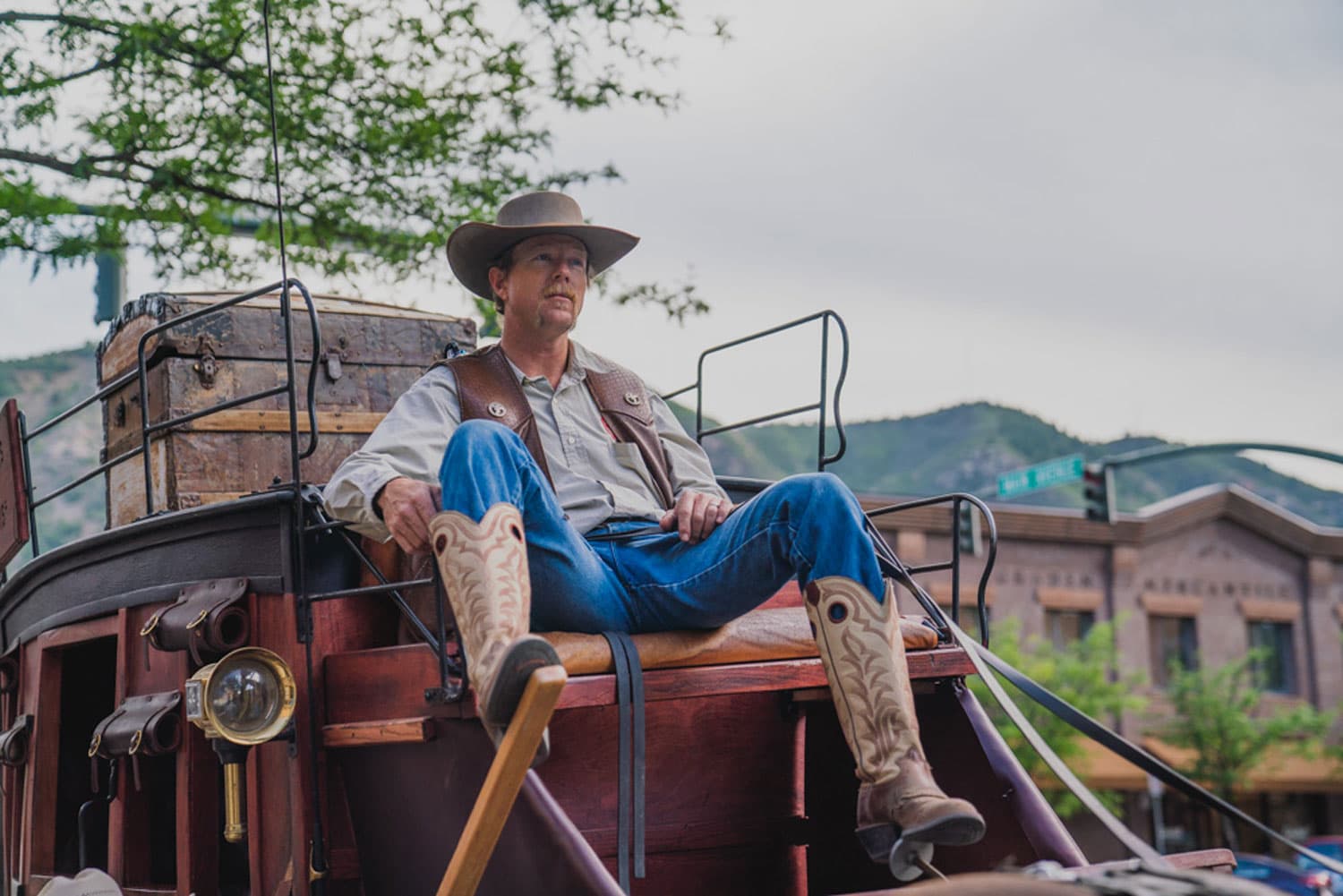 A person in cowboy boots, jeans, a leather vest and cowboy hat sits on an old-fashioned cowboy carriage in Durango, holding its reigns.
