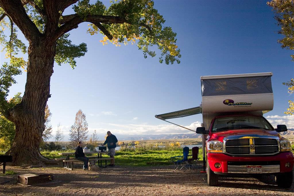 A dusty-blue summer sky glows with the warmth of the sun above a couple grilling something on a camp grill next to their red RV camper. They are in Colorado's Highlight State Park near Fruita.