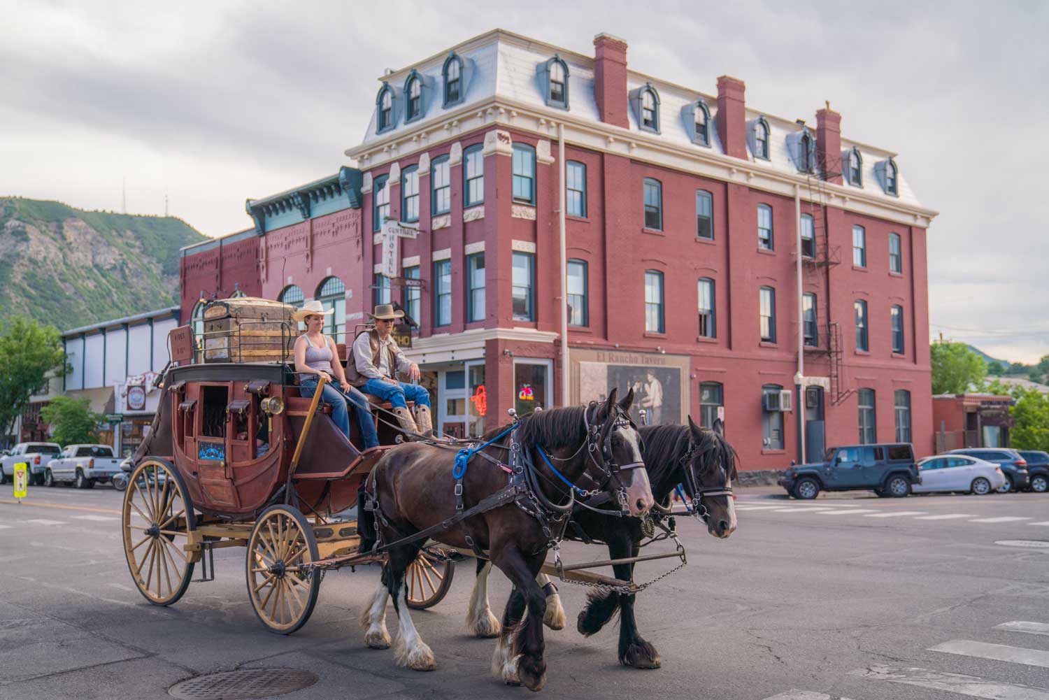 The Historic Strater Hotel sits on a corner of Durango. In front of the hotel rides by a horse-drawn carriage carried by two brown horses.