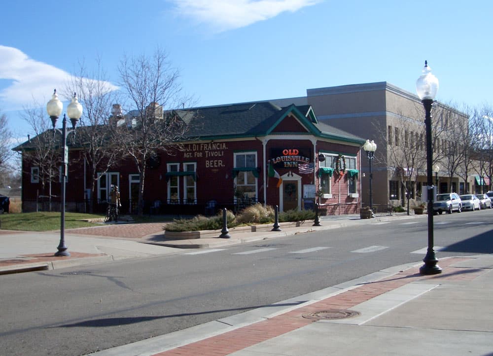 A view of the Old Louisville Inn from across the paved street. It's a red building with a green roof and a corner entrance. There's a two-story building next door and leafless trees line the sidewalk.