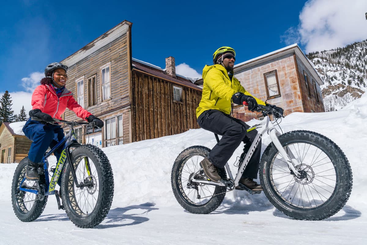 Two people wearing helmets and light winter jackets ride fat-tire bikes through the snowy streets of St. Elmo, a ghost town near Salida, Colorado.