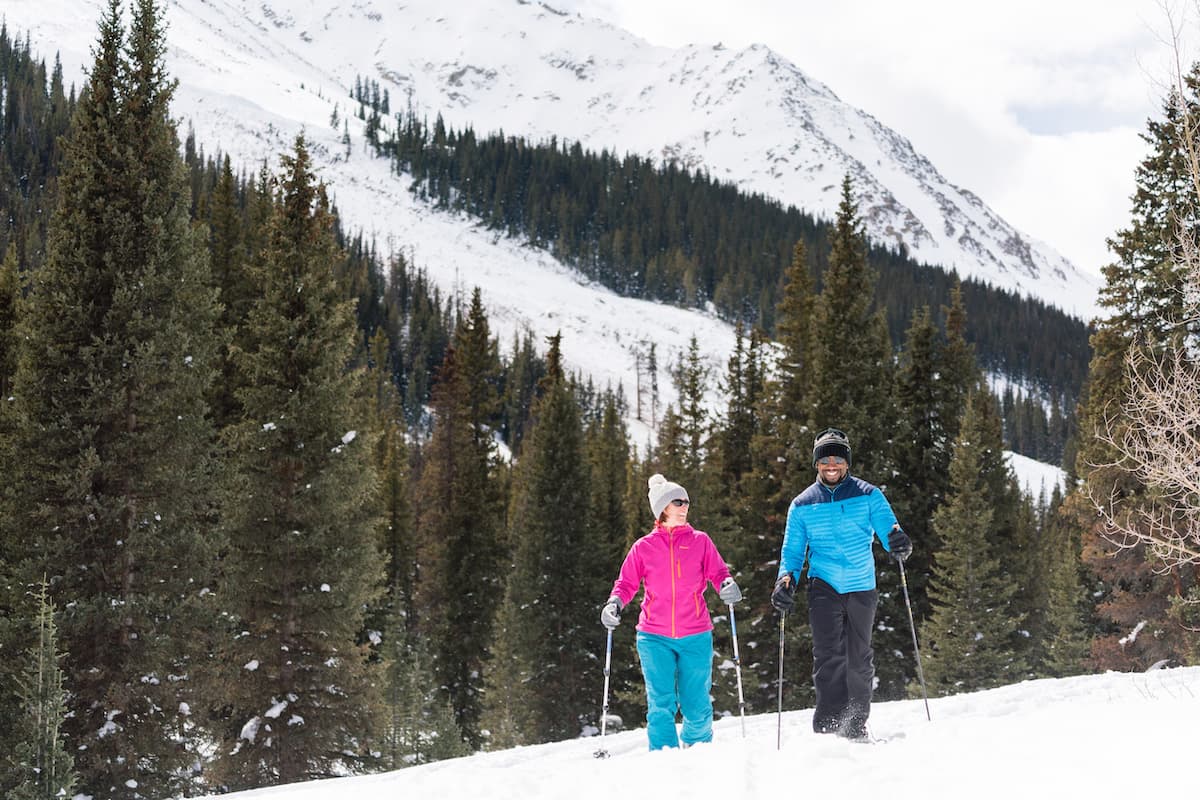 Two snowshoers in bright blues and pinks use ski poles to propel through the deep powder, surrounded by an evergreen forest