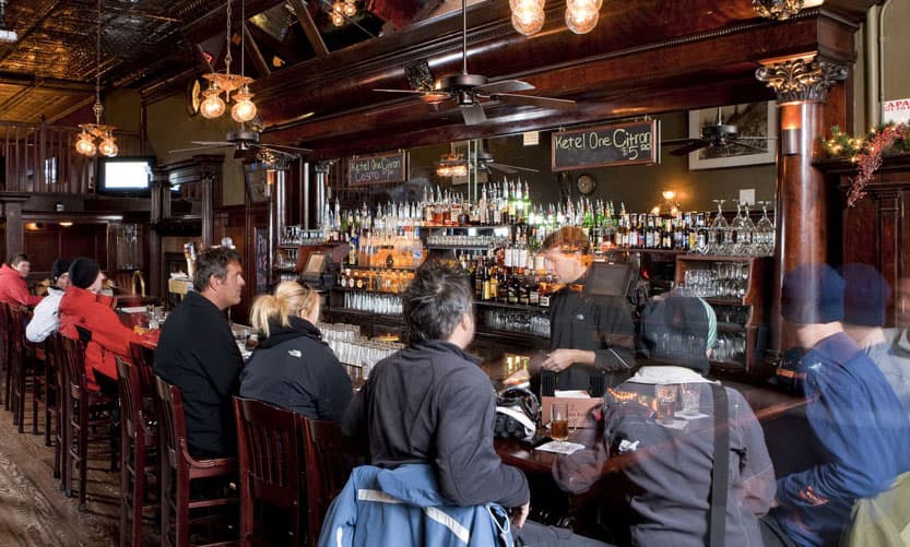 People sit in tall, dark cherry wooden chairs around a dark-wood bar in Telluride, Colorado. Many of the guests are dressed in knit hats and winter coats.