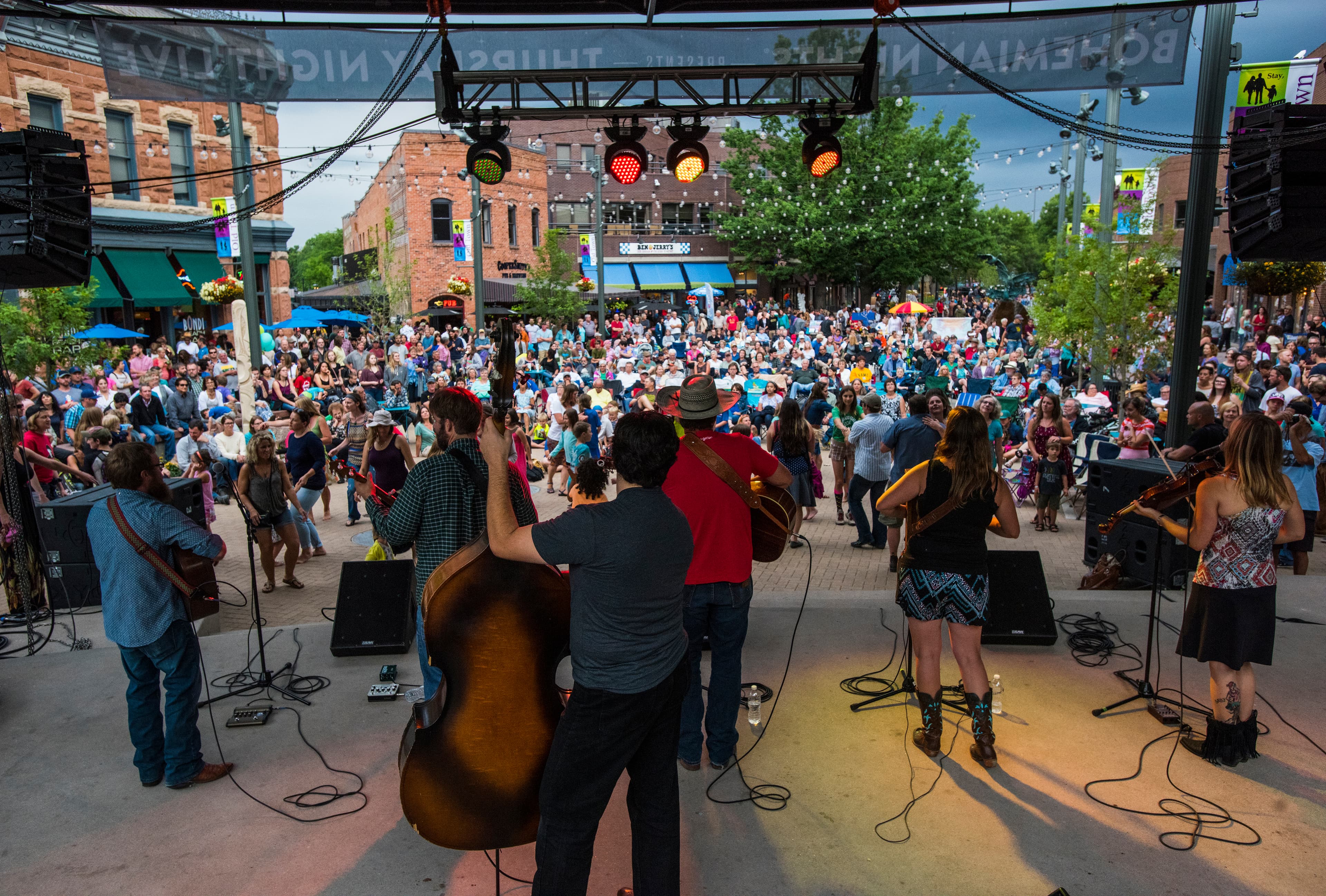 A six-person band with their backs to the camera face a large crowd outside at Old Town Square. There are twinkle lights strung up between green trees.