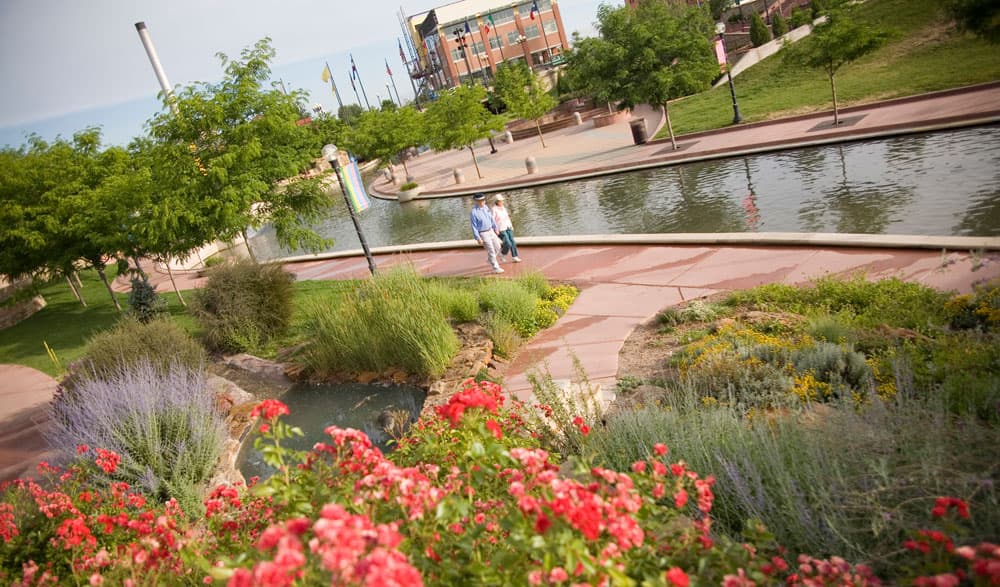 A couple walk hand in hand down a path of wide red-stone slabs that follows part of the Arkansas River in Pueblo, Colorado. Next to the path is a greenspace featuring young trees, lavender and red flowers.