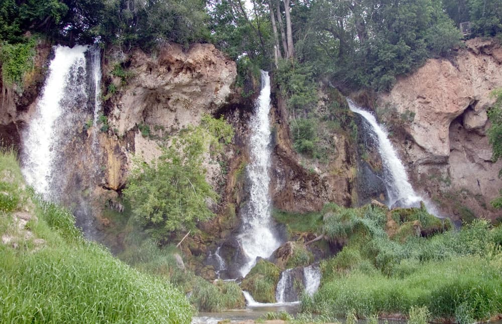 Rushing water cascades down from a rocky cliff face with lush foliage on top. The water forms three waterfalls at Rifle Falls in Colorado.