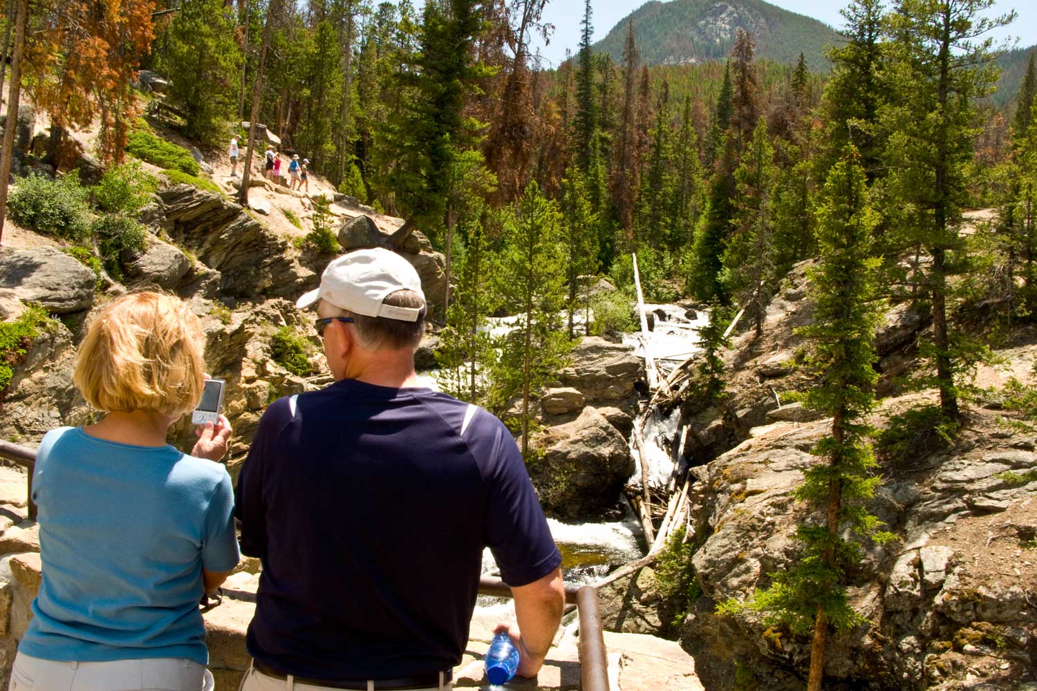 Two people sit next to each other and look out at the Adams Falls from the Rocky Mountain National Park trail. There is a small ravine with the water trickling down between tall pine trees.