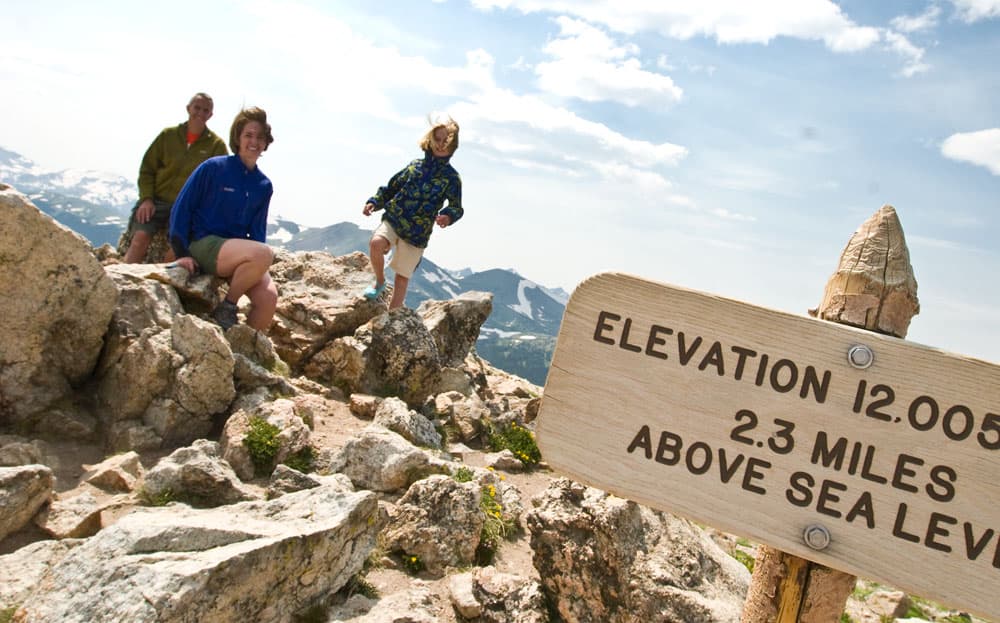 From a titled angle, we see a sign that reads "Elevation 12,005. 2.3 miles above sea level." Behind the sign, a family poses on a rocky outcrop. Behind the family, we see distant mountains dusted in snow.