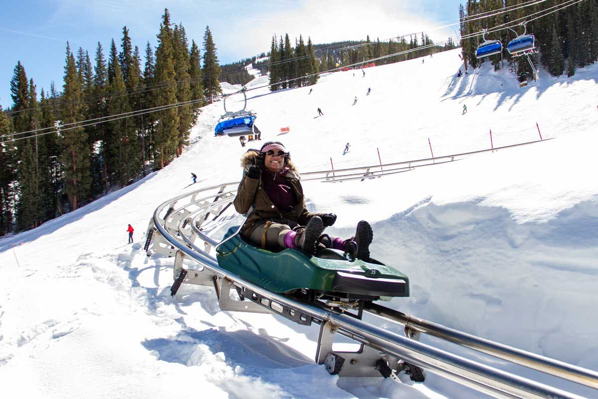 A smiling patron gives the camera the peace sign with their gloved fingers as they slide down the metal track of Copper Mountain's Rocky Mountain Coaster. There is snow piled high on the hills around the person as people ski behind them and the sun shines brightly down.
