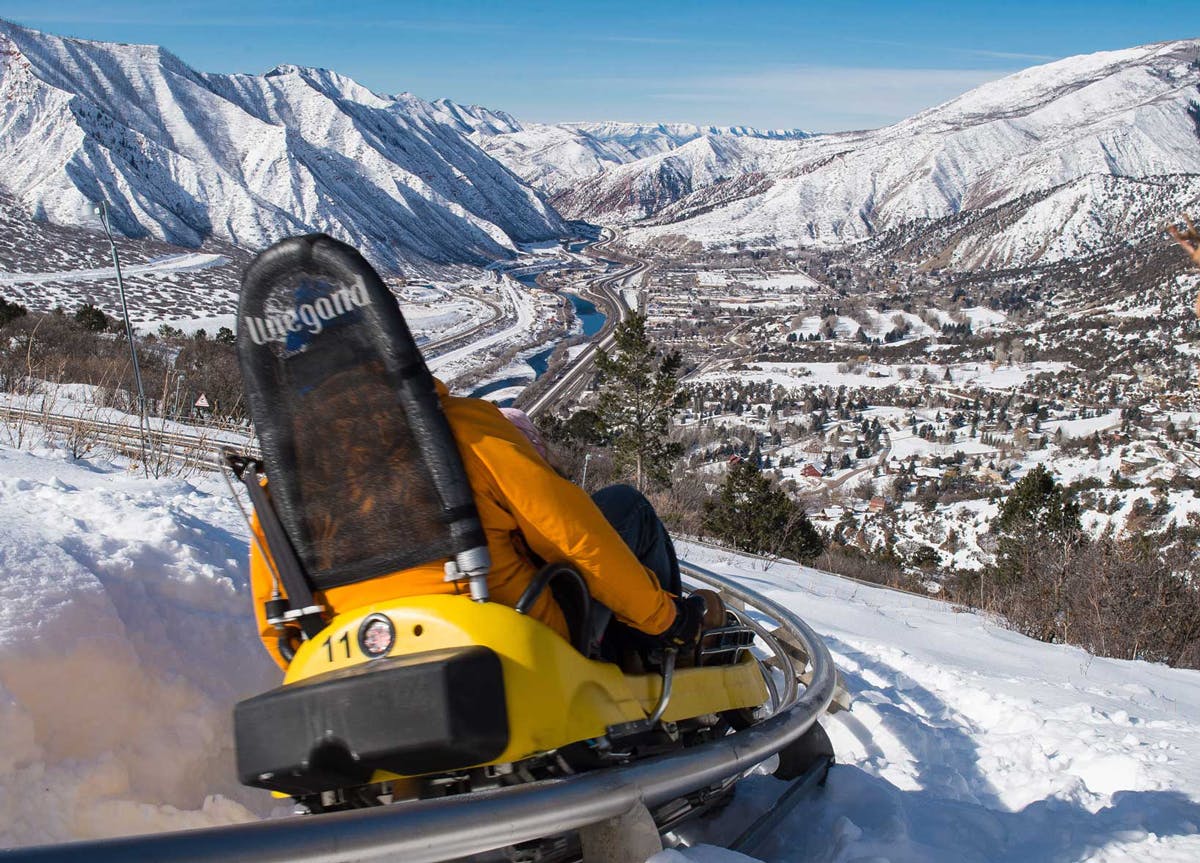 A person dressed in warm winter clothes sits on a yellow alpine coaster seat and glides down curved metal tracks on the Glenwood Caverns Adventure Park alpine coaster. In the background past the coaster seat are snow-covered jutted mountains under a blue sky.
