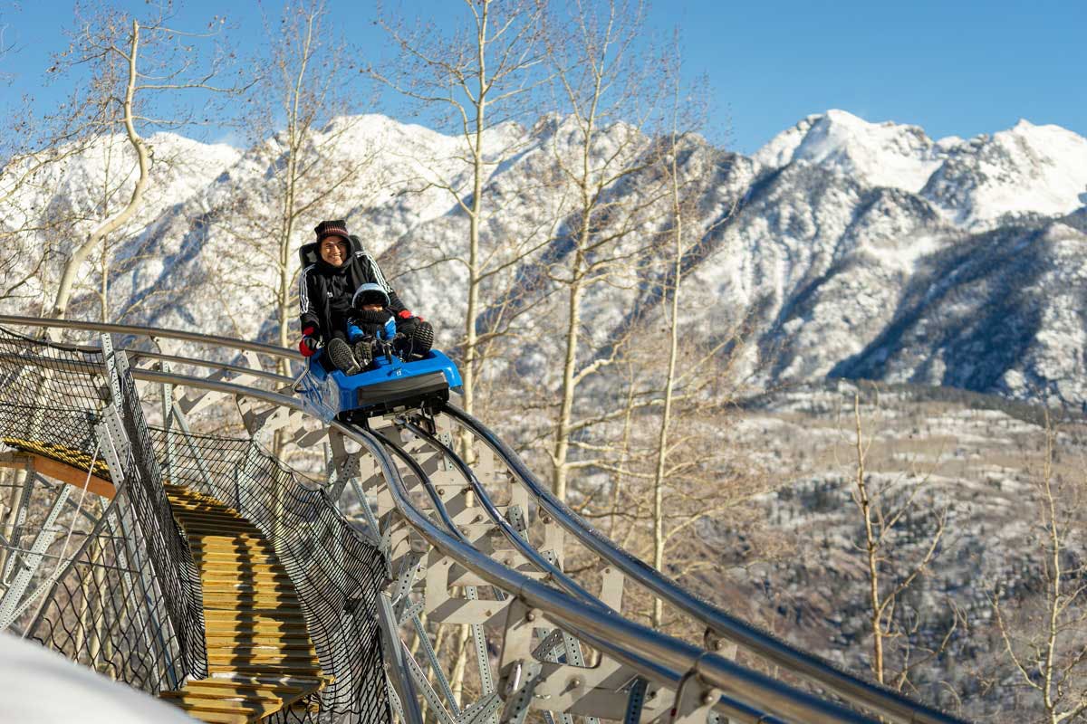A parent and their children zoom down the Purgatory Ski Resort's Mountain Coaster in Durango, Colorado, with a horizon of snowy mountain peaks in the background under a dusty-blue sky.