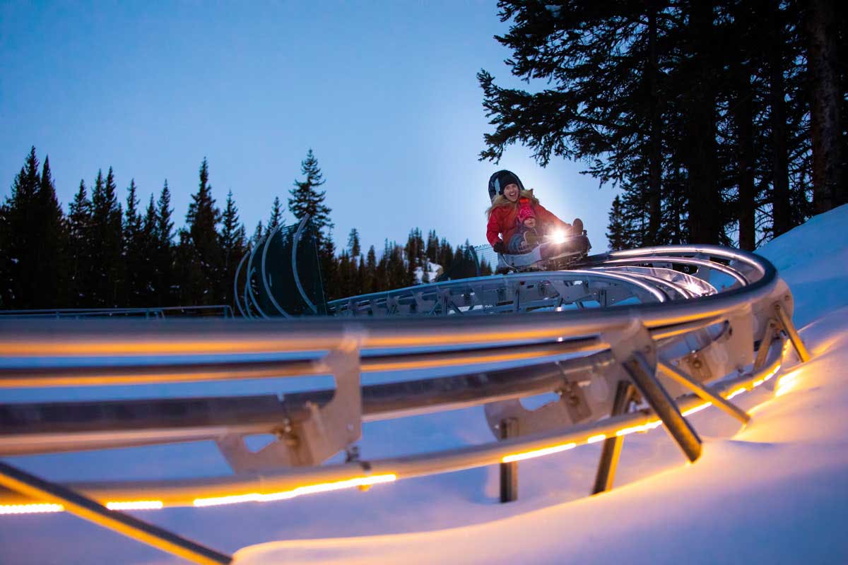 A parent and their child smile widely as they race down the metal tracks of the Aspen Snowmass' Breathtaker Alpine Coaster. The sun has gone down and left a hint of blue in the sky but the tracks are lit up with white light that glows off the snow beneath the track.